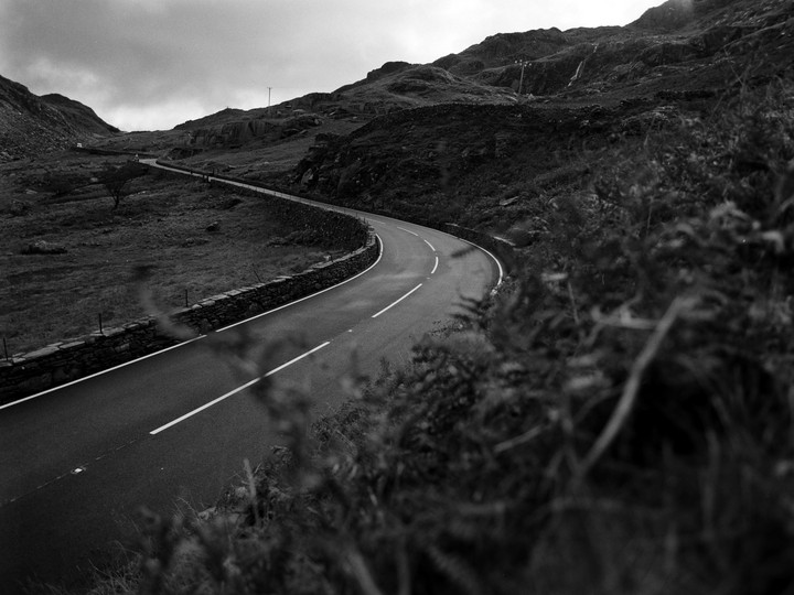 A winding road with a stone wall on the left leads through a mountainous landscape under a cloudy sky.