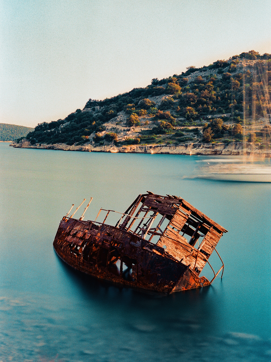 A rusted shipwreck sits in calm blue water with a green, tree-covered hill in the background.