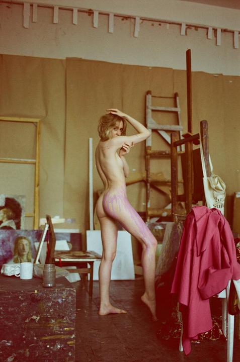 A nude woman with purple paint on her body stands in an art studio.