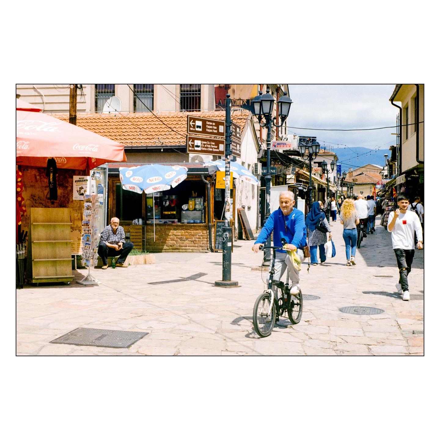 A man rides a bicycle down a busy street with shops and people.