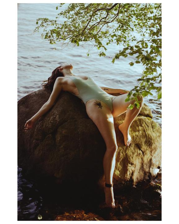 A woman in a swimsuit lies on a large rock at the edge of a lake with tree branches overhead.