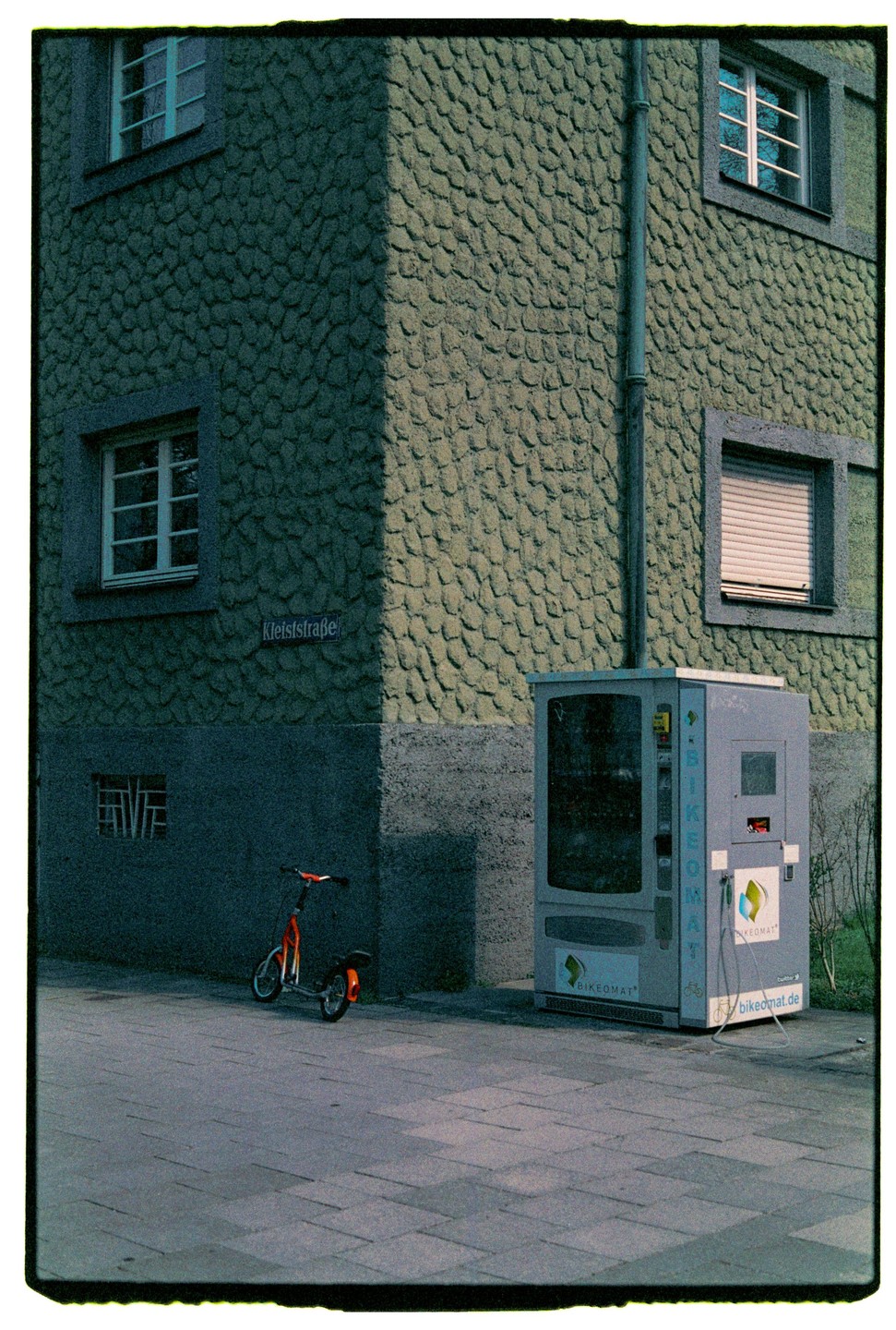A scooter is parked on a sidewalk next to a building with a textured facade and a bikeomat machine.