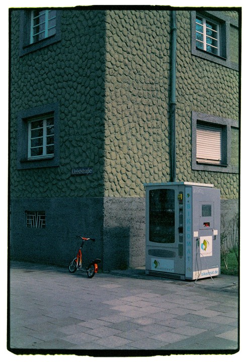 A scooter is parked on a sidewalk next to a building with a textured facade and a bikeomat machine.