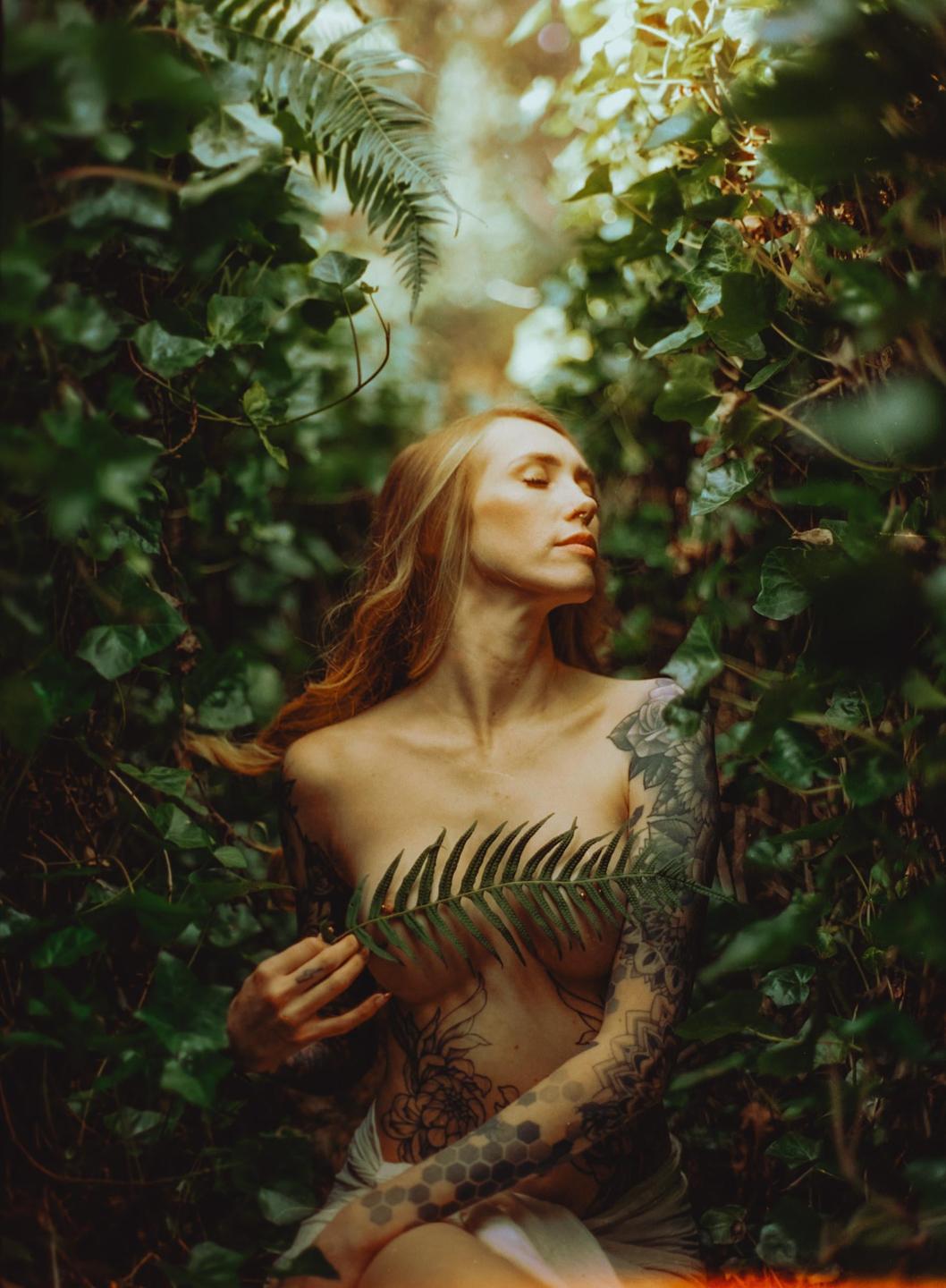 A woman with long hair and tattoos is partially covered by a fern leaf in a lush green setting.