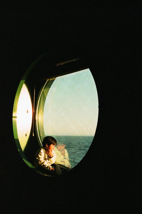 A child sits by a round window looking out at the ocean, with a reflection of another child visible in the glass.