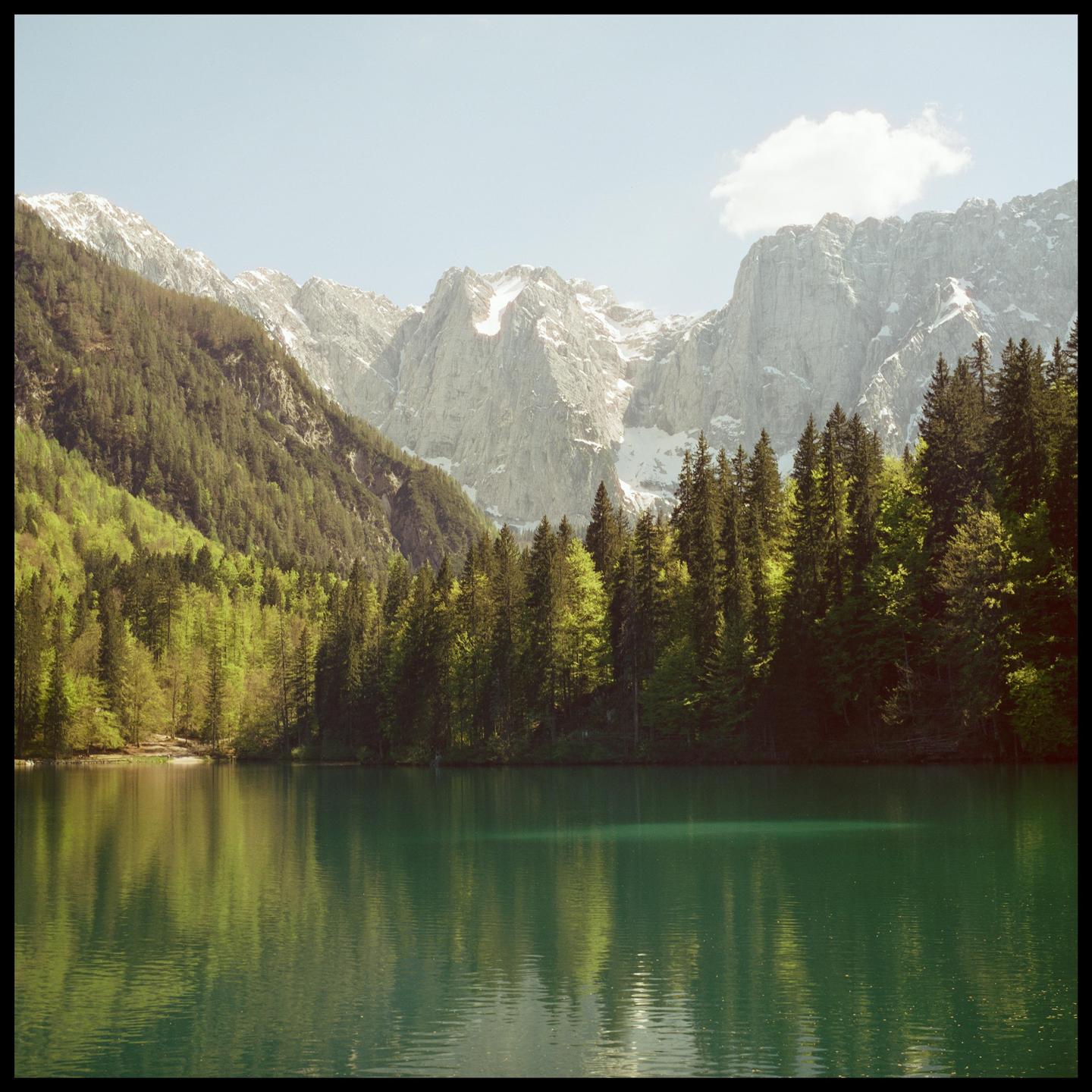 A lake with green water reflects a forest and snow-capped mountains under a clear sky.