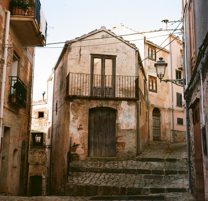 A narrow, cobbled street with stone steps leads up to old, weathered buildings with balconies and wooden doors.