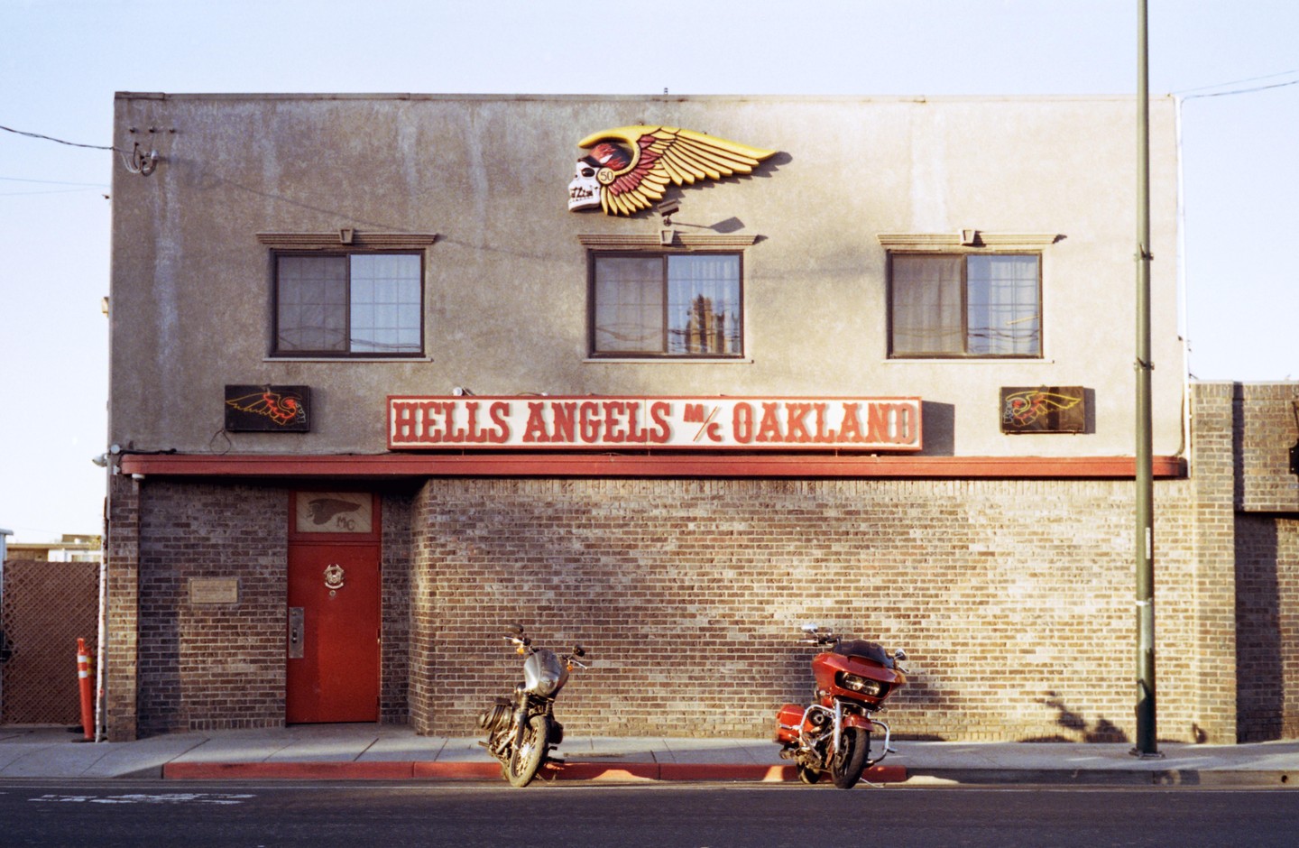 Two motorcycles are parked in front of a brick building with a "Hells Angels MC Oakland" sign and a skull with wings logo.