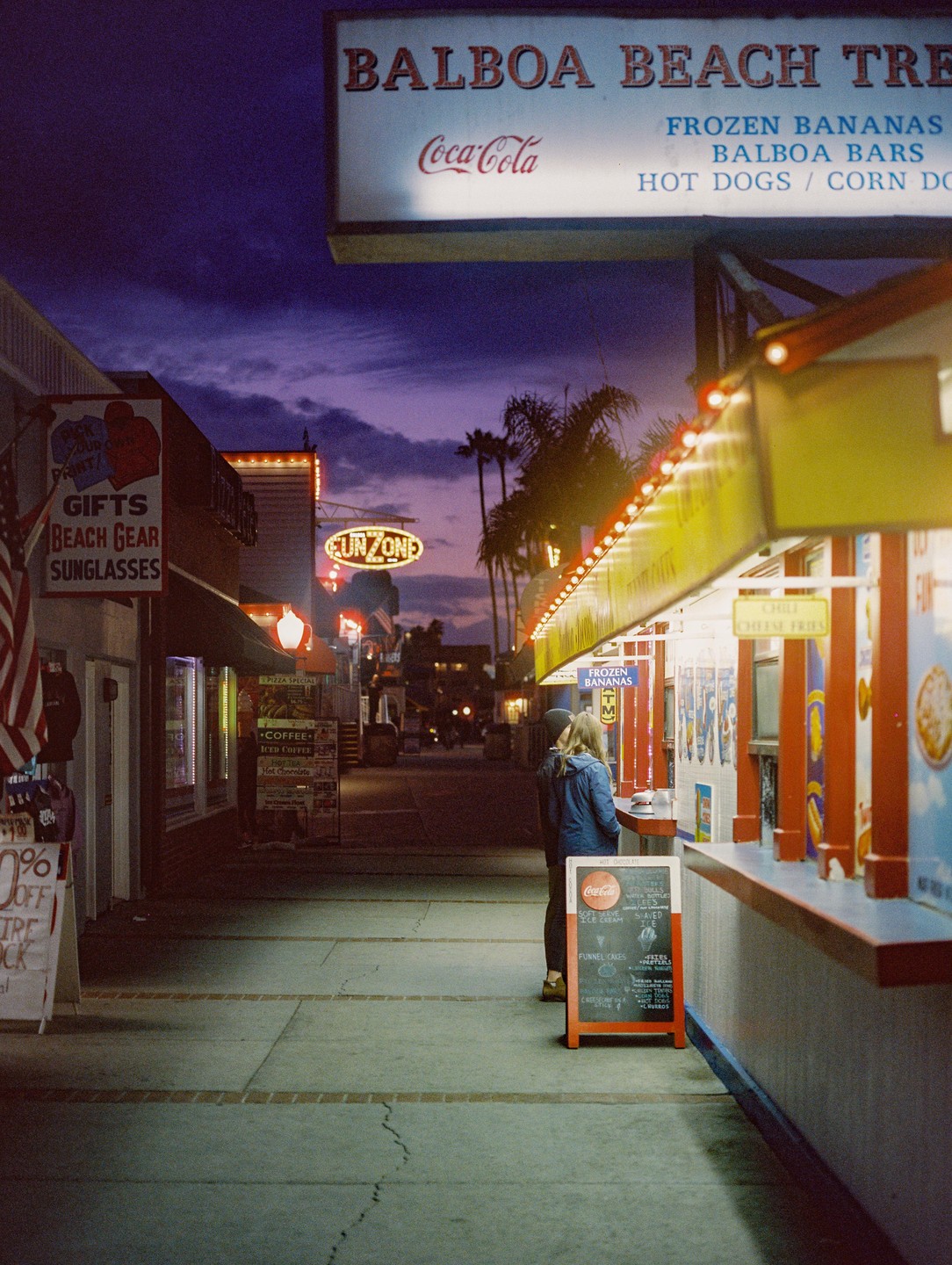 A woman stands at a food stall on Balboa Beach at dusk.