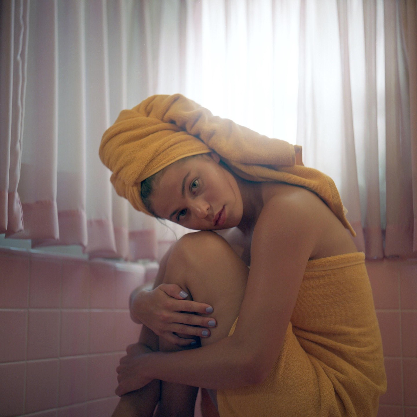 A woman wrapped in a yellow towel sits on the floor of a pink tiled bathroom.
