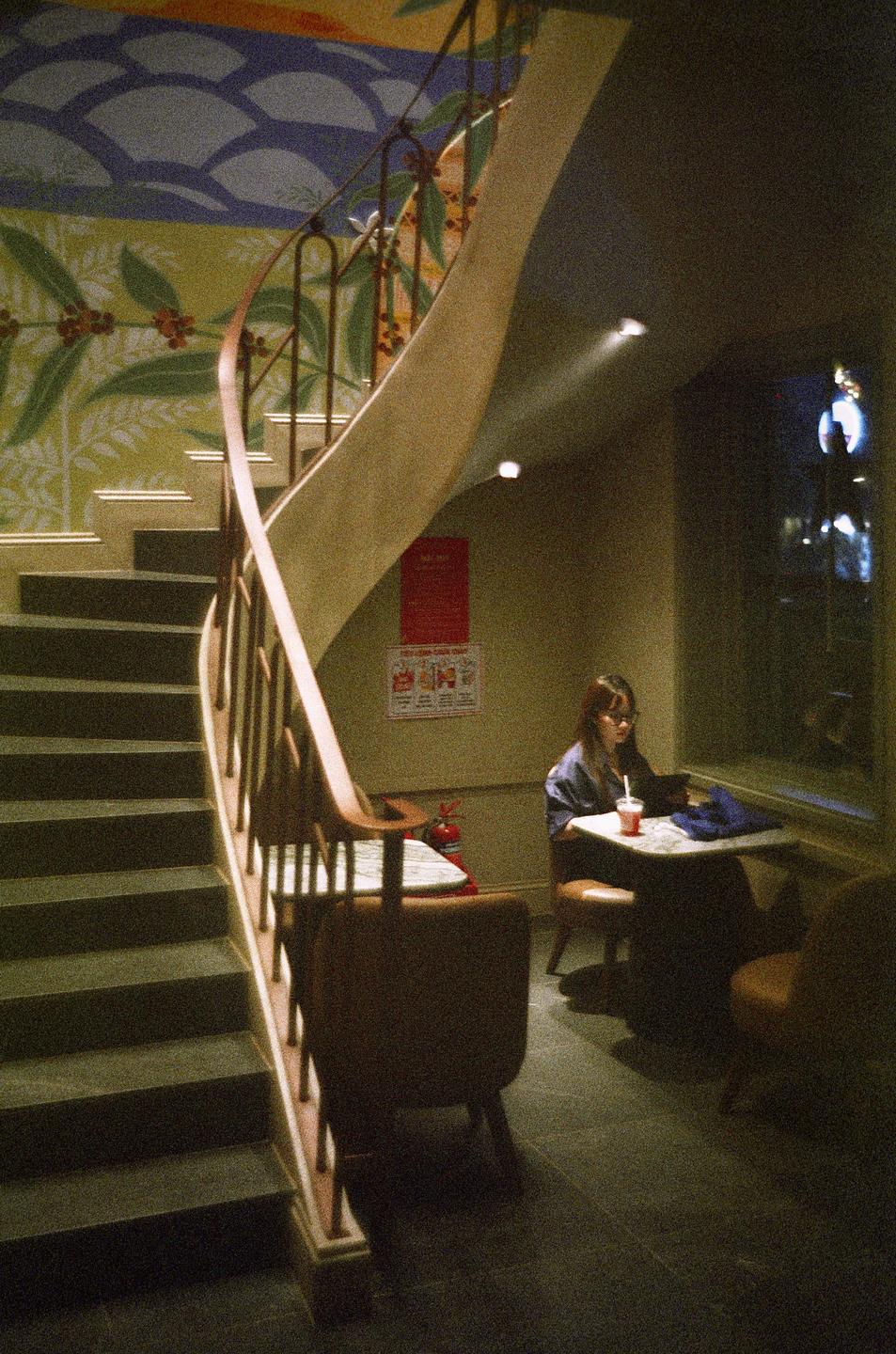A woman sits at a table next to a window and a spiral staircase in a cafe.