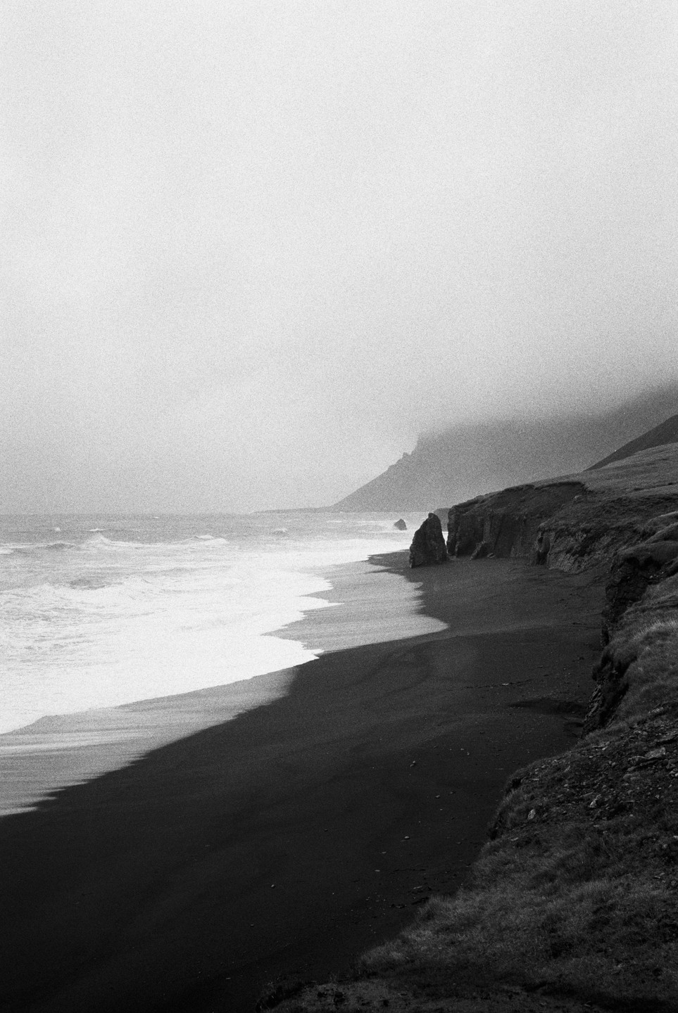 Waves crash on a black sand beach next to a cliff under a hazy sky.