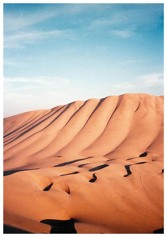 A vast desert landscape shows sand dunes with long shadows under a clear sky.