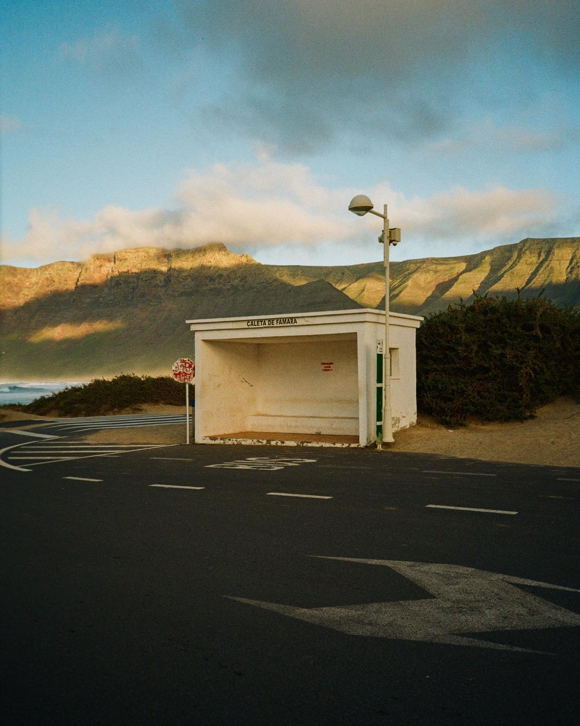 A bus stop with a street light stands beside a road with mountains and the ocean in the background.