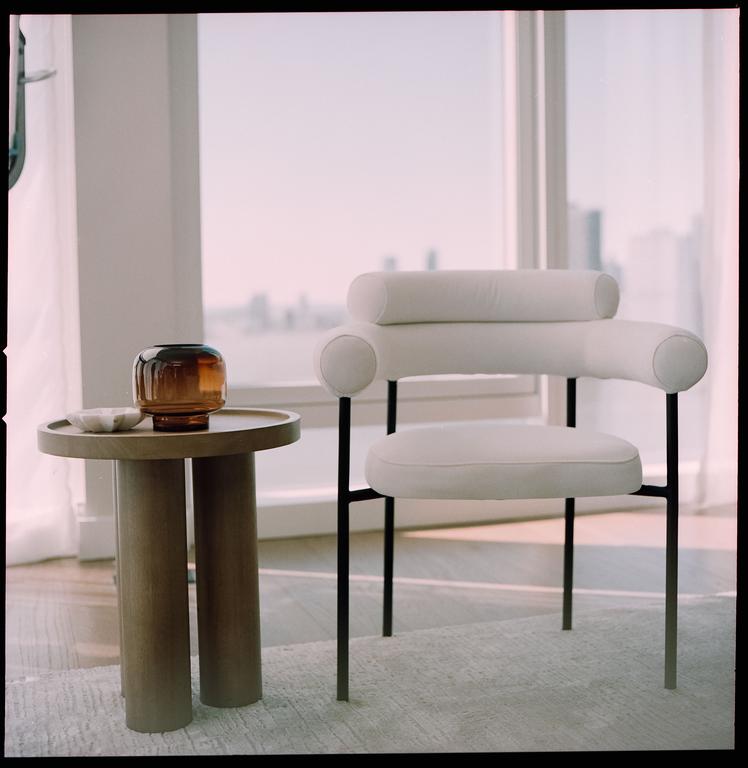 A modern white chair and a round side table with a brown vase are positioned in front of a large window overlooking a city.