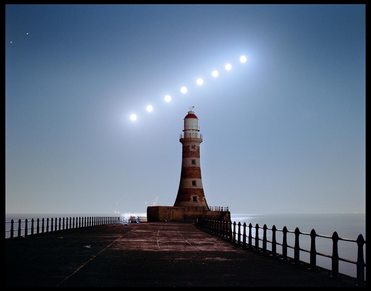 A lighthouse stands on a pier with a sequence of moons rising in the sky.