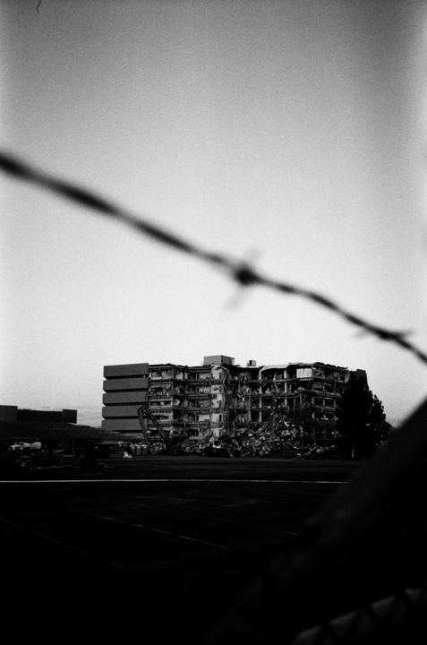A demolished building is visible behind a blurred barbed wire fence in the foreground.