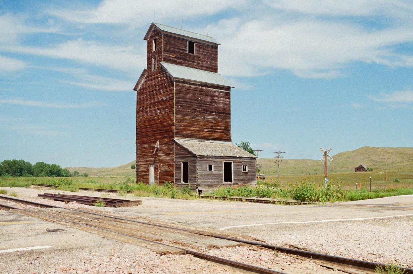An abandoned wooden grain elevator stands beside railroad tracks in a rural landscape under a blue sky with clouds.