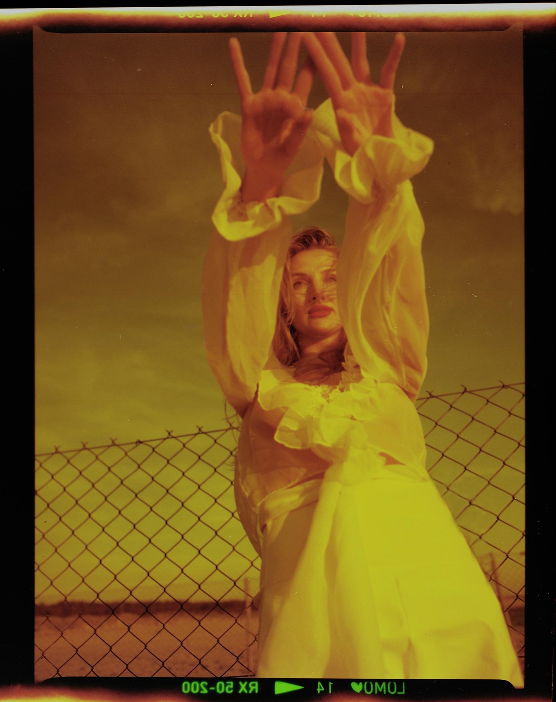 A woman in a flowing yellow dress raises her hands in front of a chain-link fence under a yellow sky.
