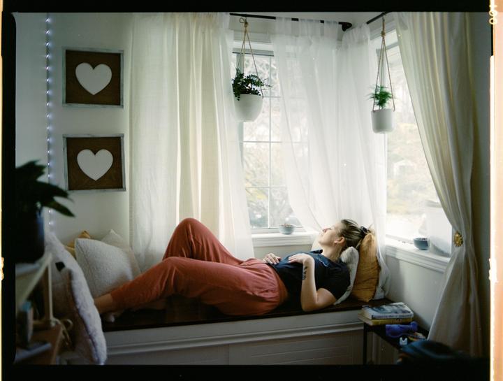 A woman lies on a window seat with sheer white curtains and hanging plants.