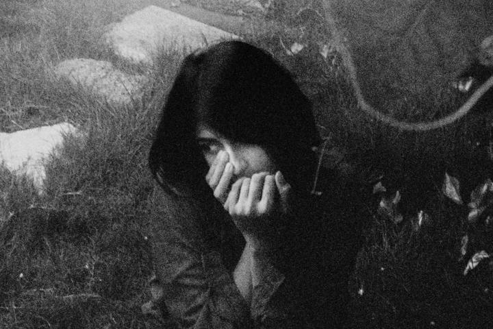 A woman with dark hair covers her mouth with her hands while looking towards the viewer.