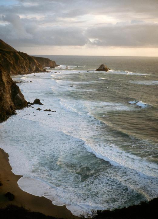 Waves crash against a rocky coastline under a cloudy sky.