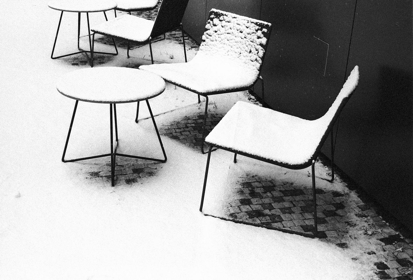 Several outdoor chairs and tables are covered in a fresh layer of snow next to a dark wall.