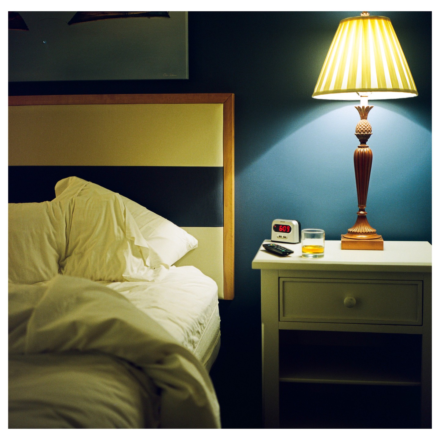 A bedside table with a lamp, alarm clock, remote control, and a glass of amber liquid sits next to an unmade bed.