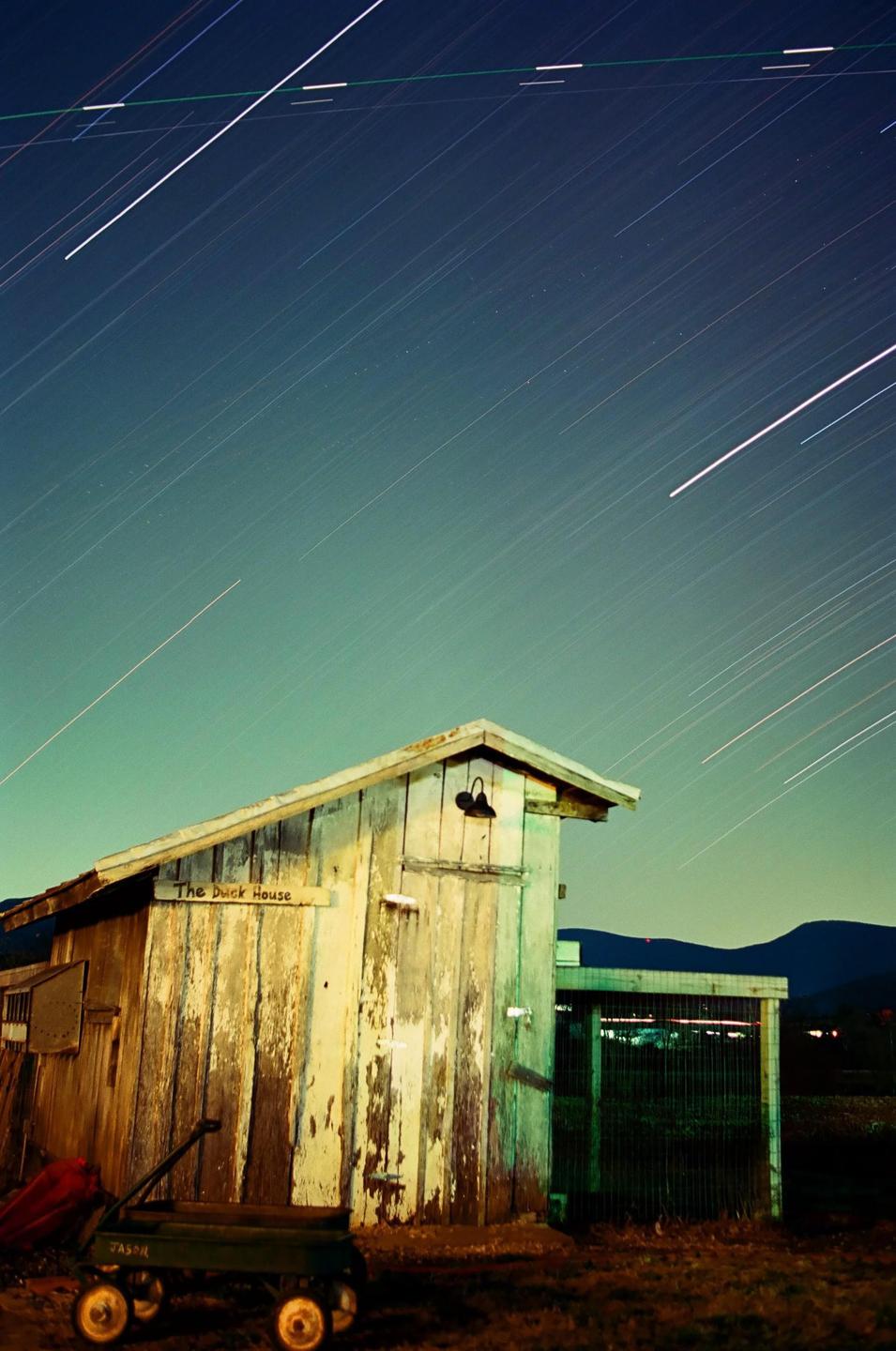 A wooden shed with peeling paint stands under a night sky filled with star trails.