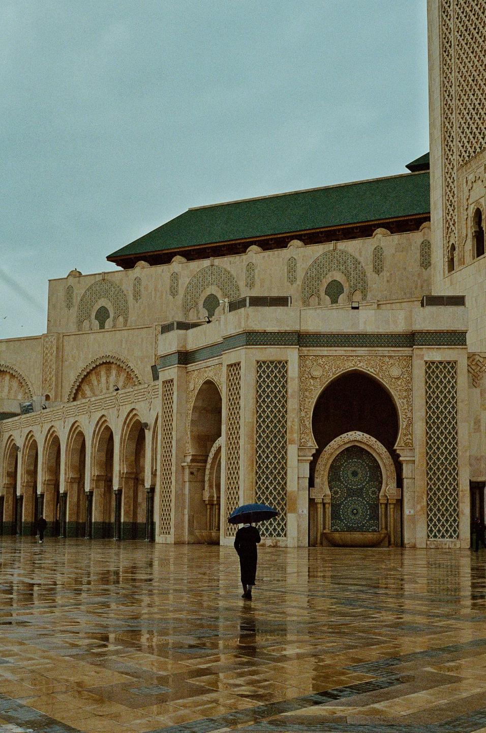 A person holding an umbrella walks across a wet, tiled courtyard towards a large, ornate building with a green roof.