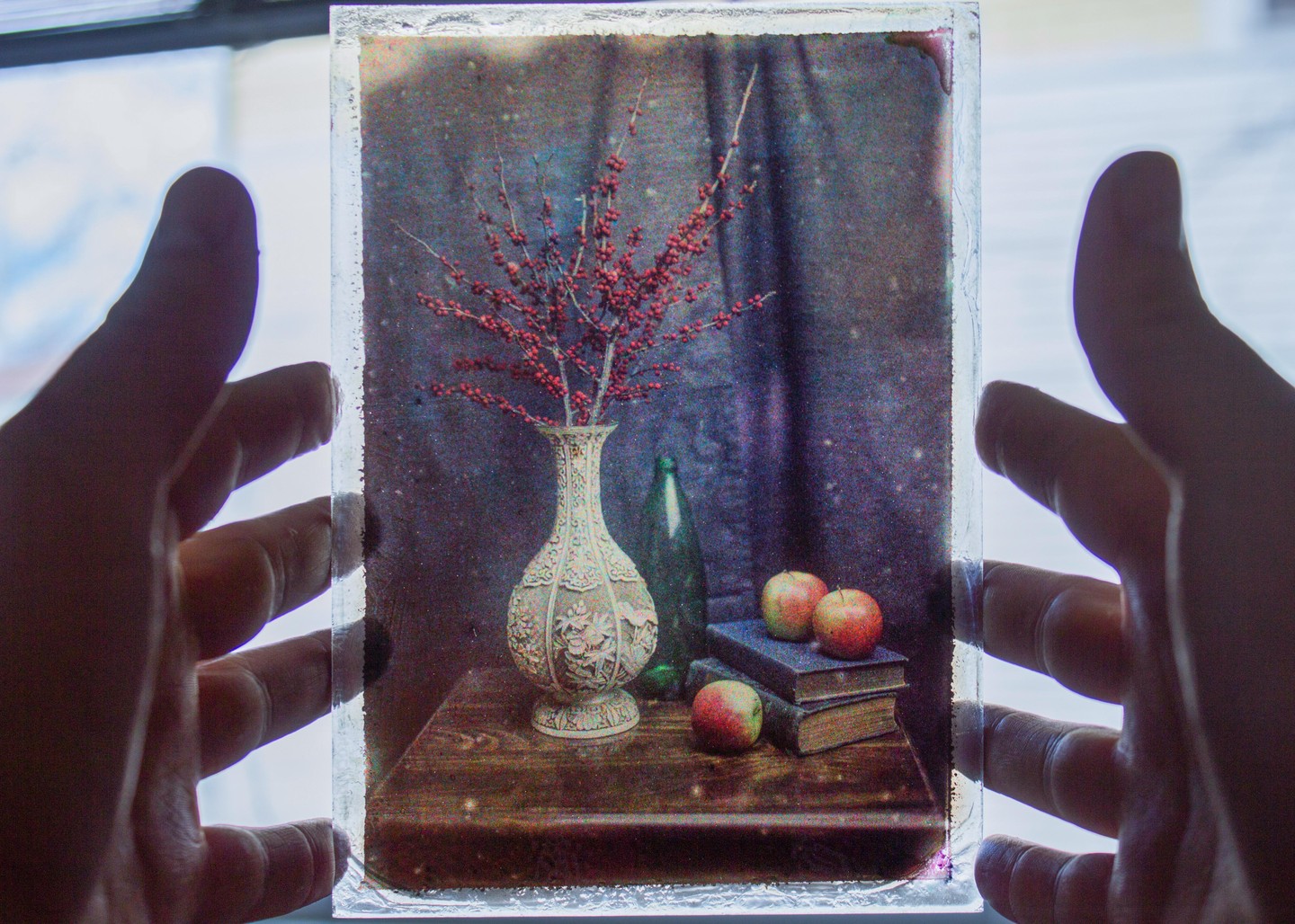 A still life of juniper berries in a vase, a bottle, apples, and books is held up to the light by two hands.