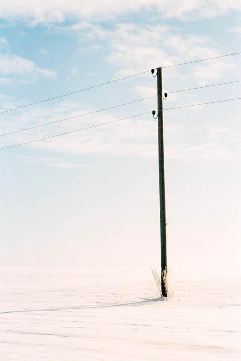A lone utility pole stands in a vast, snow-covered field under a light blue sky with wispy clouds.