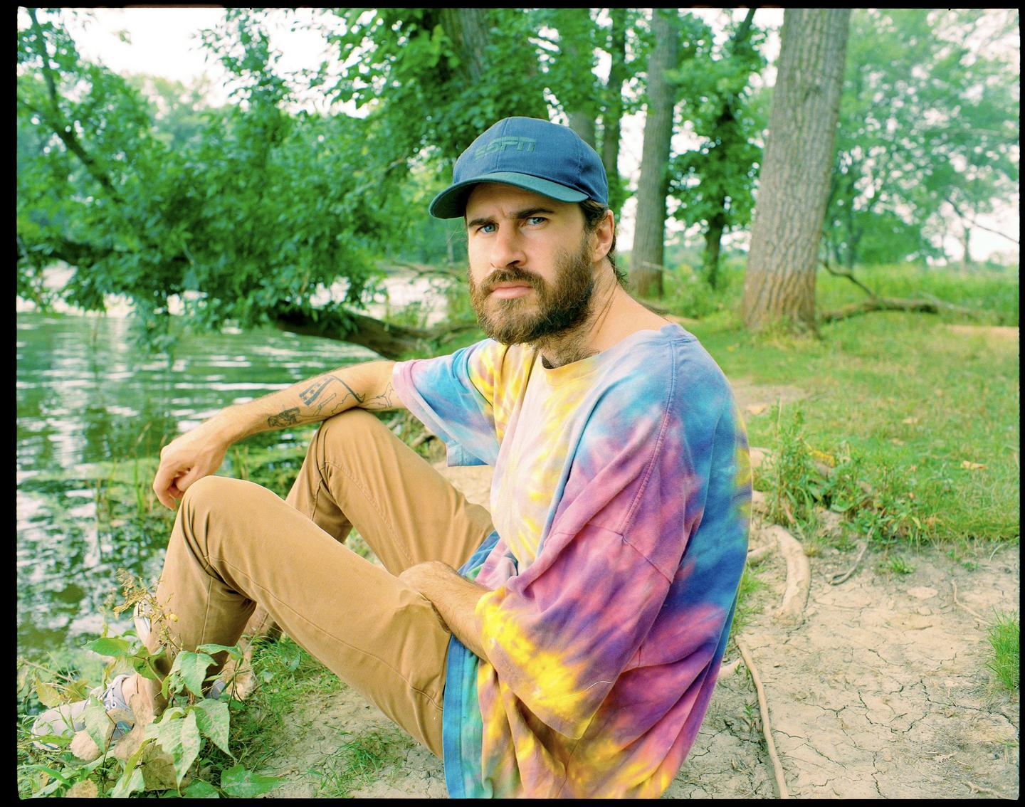 A man with a beard and a tie-dye shirt sits by a body of water with trees in the background.