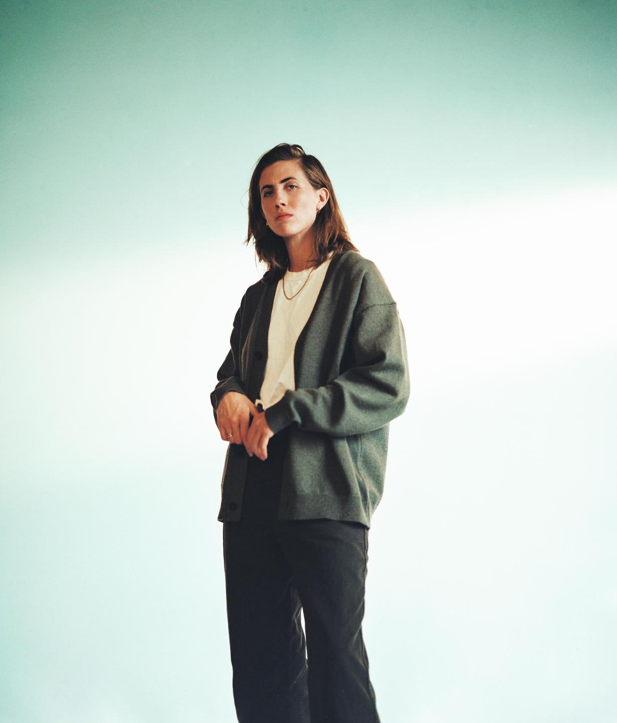 A woman with dark hair stands against a light blue background, looking directly at the viewer.