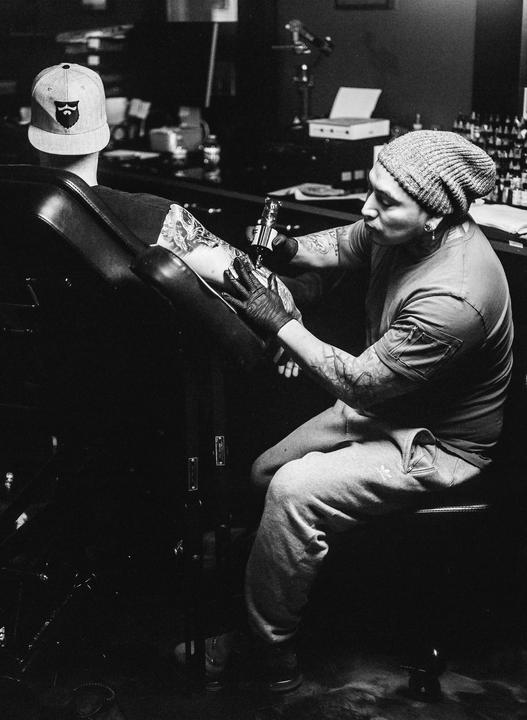 A man with a beanie and tattooed arms is tattooing the arm of another man wearing a baseball cap.