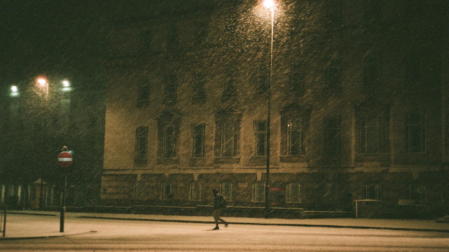 A person walks across a snow-covered street at night, illuminated by a street light, with a building in the background.