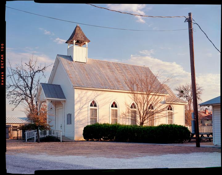 A white wooden church with a bell tower stands under a blue sky with scattered clouds.