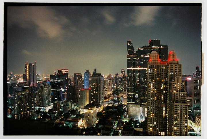 A city skyline with numerous illuminated skyscrapers is visible at night under a cloudy sky.