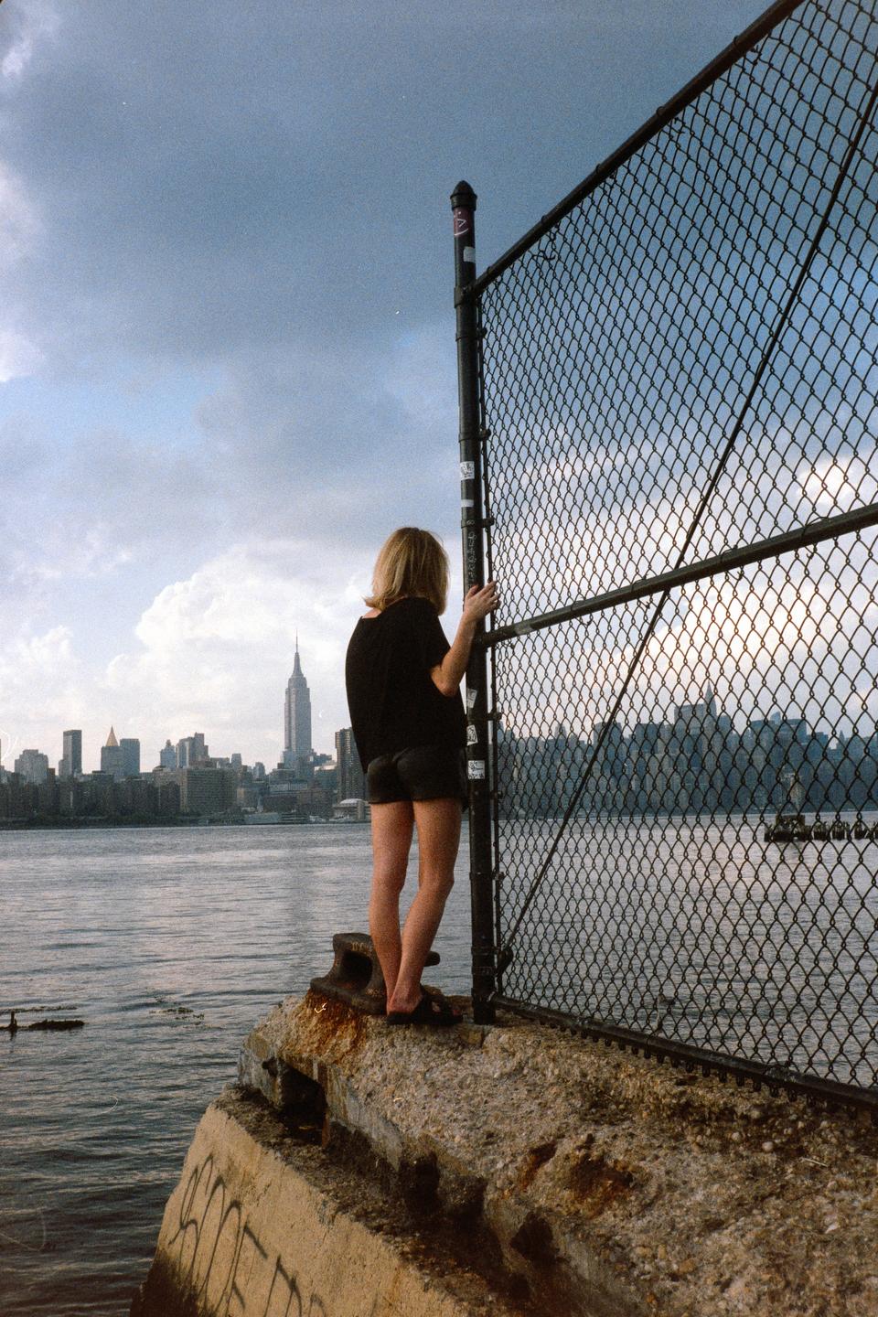 A woman stands on a concrete ledge next to a chain link fence, looking out at a city skyline across a body of water.