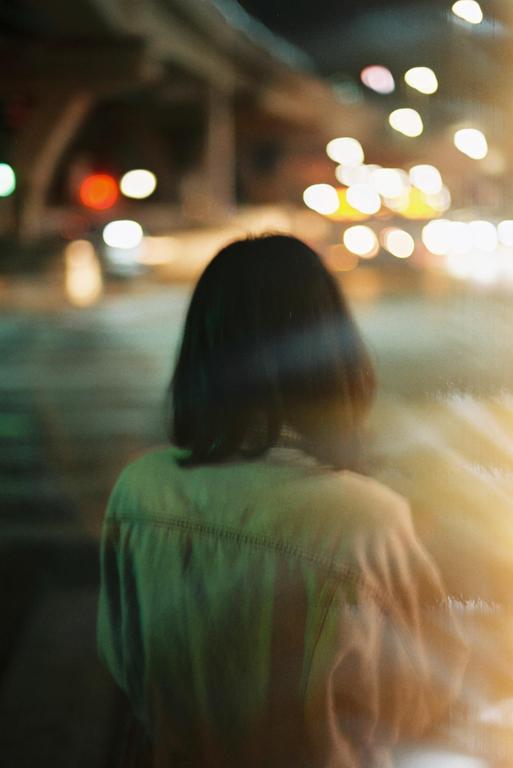 A woman with short dark hair stands with her back to the viewer, looking at blurred lights at night.