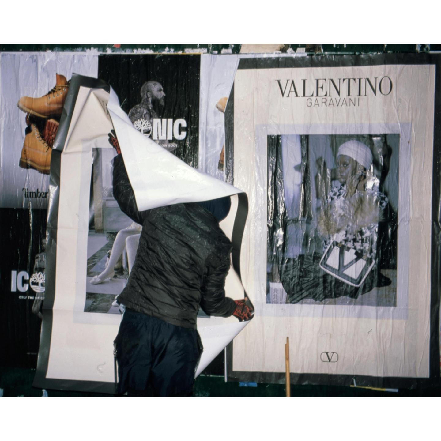 A person is peeling a large poster off a wall covered with other posters.