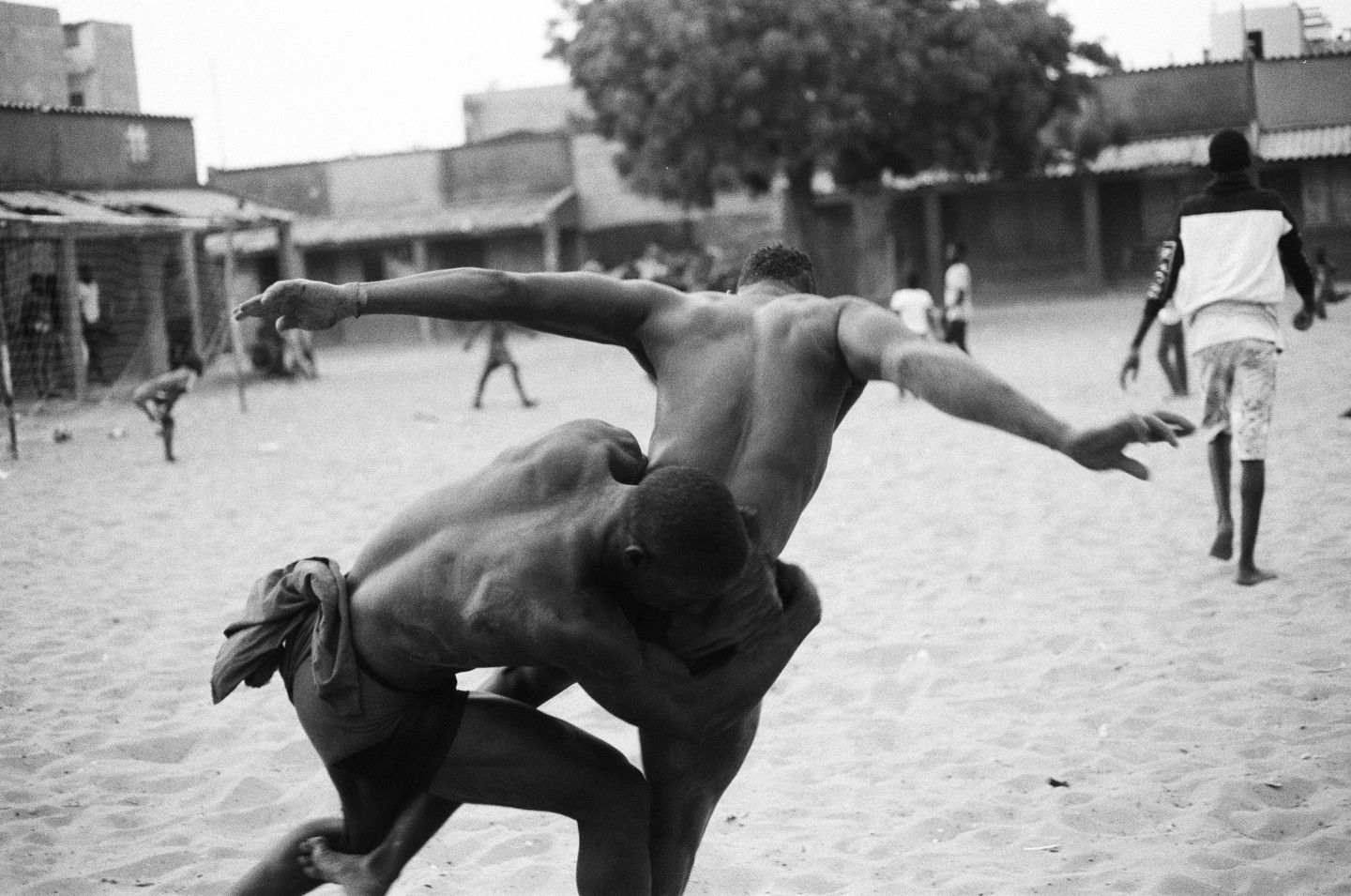 Two men wrestle on a sandy ground with buildings and a tree in the background.