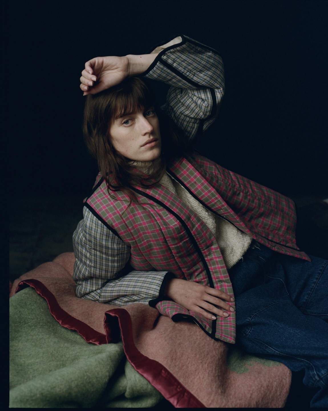 A woman with long hair and bangs lies on a blanket, wearing a plaid jacket and jeans.