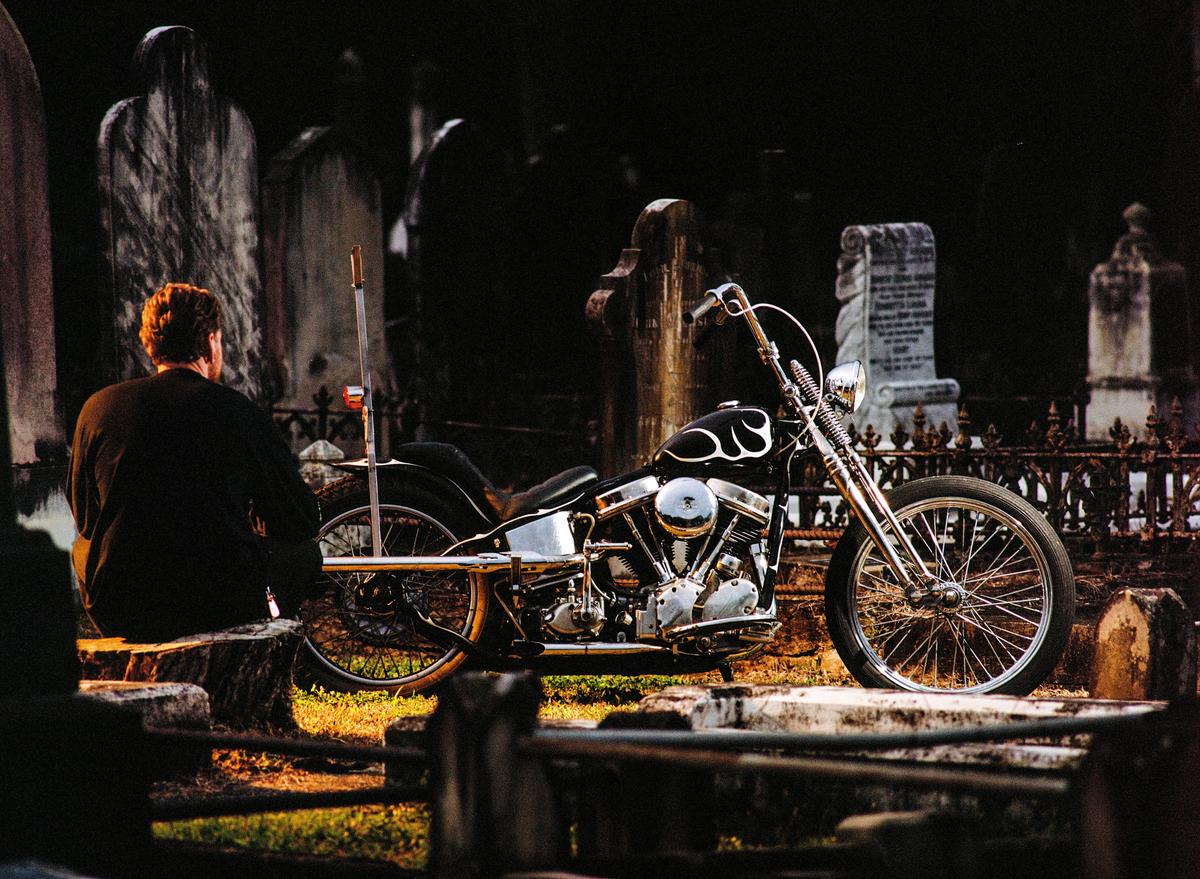 A man sits on a stone wall next to a motorcycle in a cemetery.