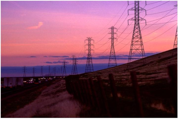 A row of power lines stands against a vibrant pink and purple sunset sky next to a highway with traffic.