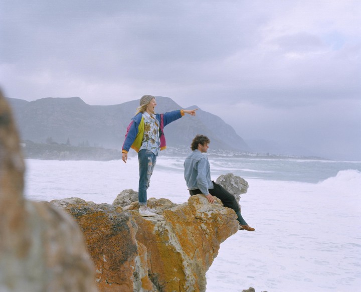 A woman points towards the ocean while standing on a rocky cliff next to a man sitting on the edge.