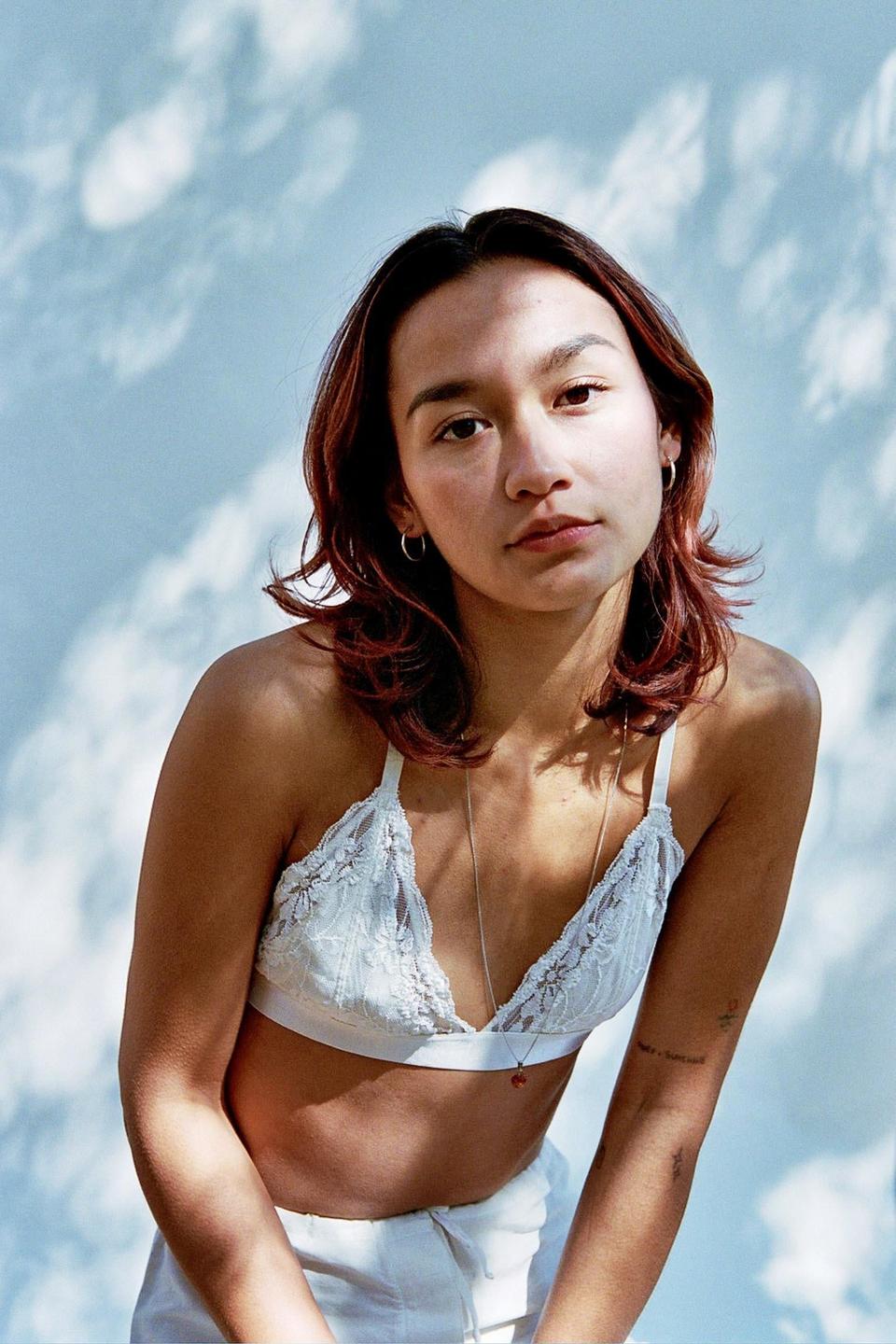 A woman wearing a white lace bralette and white pants looks directly at the viewer.