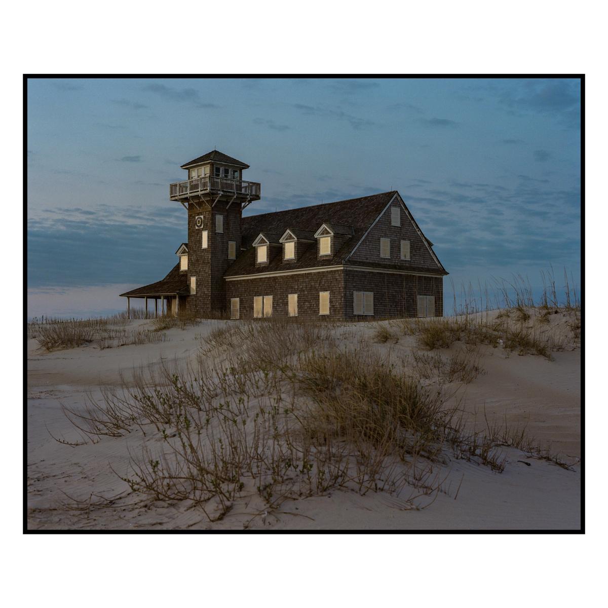 A large wooden building with a tower stands on a sandy dune under a cloudy sky.