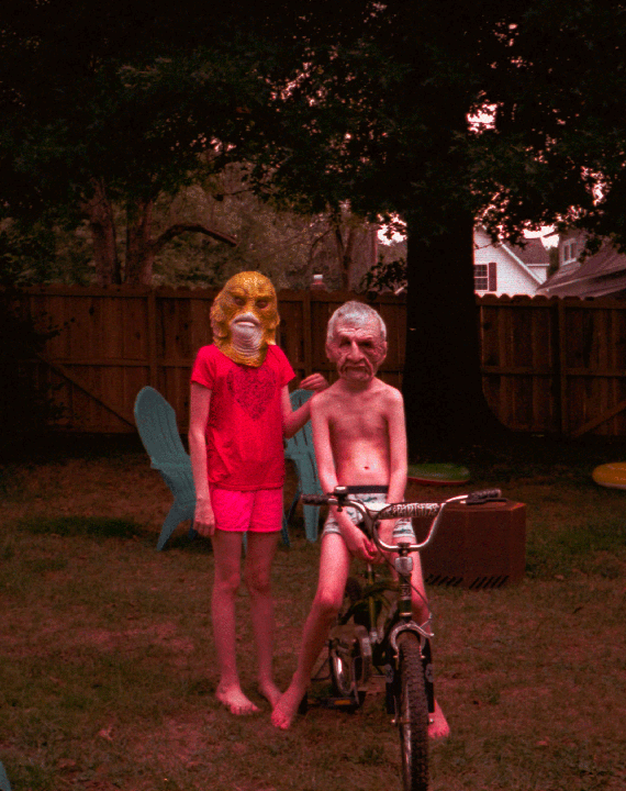 Two children wearing masks stand in a grassy backyard, one on a bicycle.
