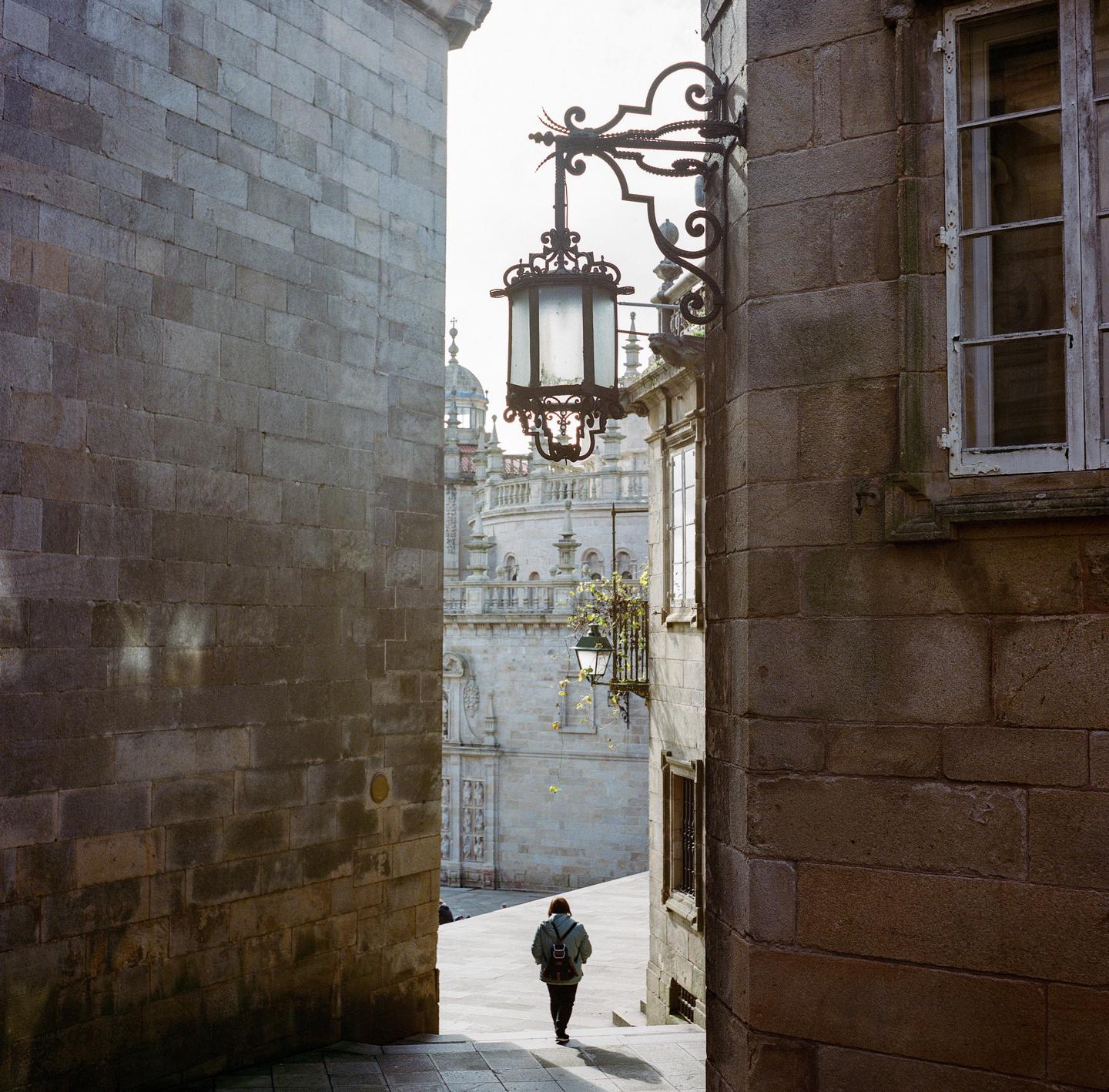 A woman walks down a narrow alleyway towards a large ornate building.